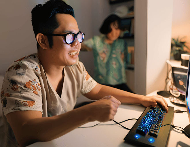 An Indonesian man in a well-lit bedroom is engrossed in his gaming PC, his expression reflecting enjoyment as he engages with the captivating visuals.